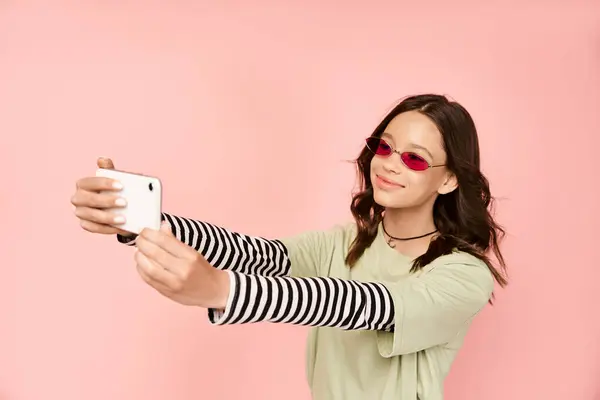 Una adolescente de moda en traje vibrante y gafas de sol tomando una selfie con un teléfono celular. - foto de stock