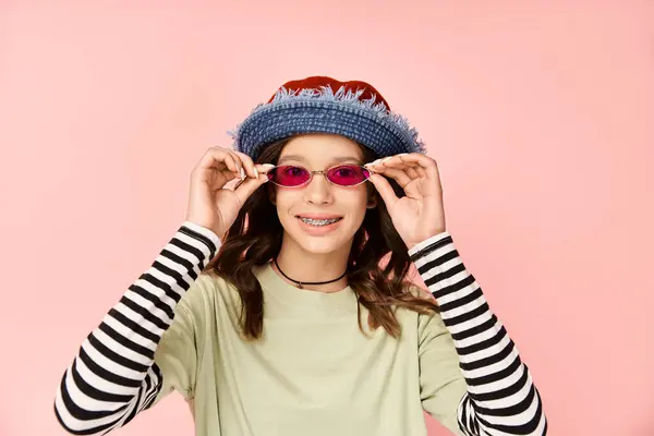 Une adolescente élégante prend une pose en tenue vibrante, portant des lunettes de soleil et un chapeau. — Photo de stock