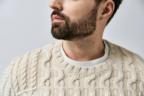 Ein stilvoller Mann mit Bart posiert in einem gemütlichen weißen Pullover vor schickem grauen Hintergrund in einem Studio. — Stockfoto