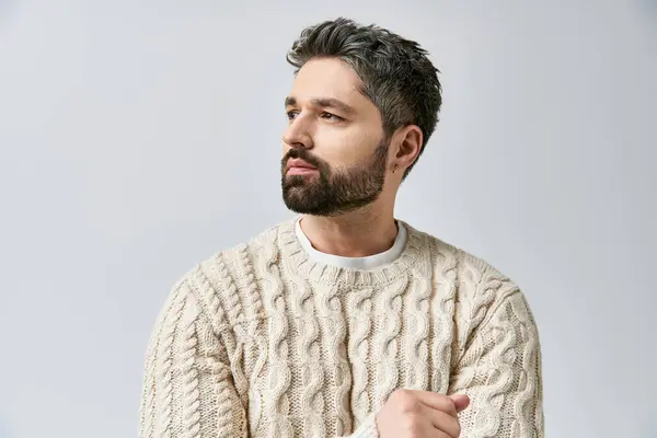 Ein charismatischer Mann mit Bart posiert im weißen Pullover vor grauem Studiohintergrund. — Stockfoto