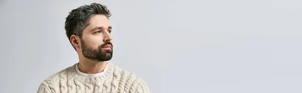 Ein charismatischer Mann mit Bart posiert in einem gemütlichen weißen Pullover vor grauem Studiohintergrund. — Stockfoto