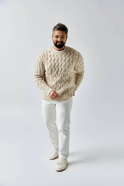 Un hombre barbudo exuda encanto con un suéter blanco y pantalones sobre un fondo gris estudio. - foto de stock