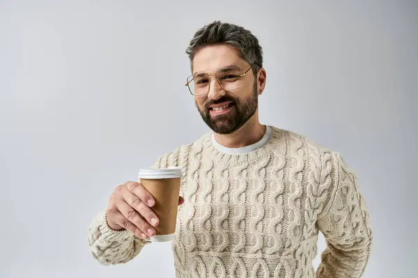 Uomo barbuto in maglione bianco tiene una tazza di caffè fumante in un accogliente ambiente monolocale su uno sfondo grigio. — Foto stock