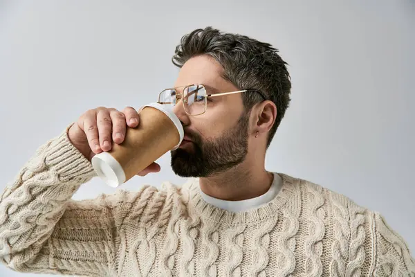 Ein bärtiger Mann im weißen Pullover, der ein Getränk aus einer Tasse genießt, während er eine Brille auf grauem Studiohintergrund trägt. — Stockfoto