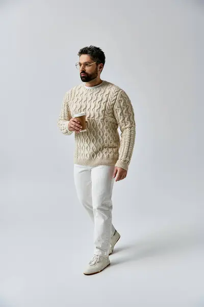 Ein bezaubernder Mann mit Bart posiert in weißem Pullover und passender Hose vor grauer Studiokulisse. — Stockfoto
