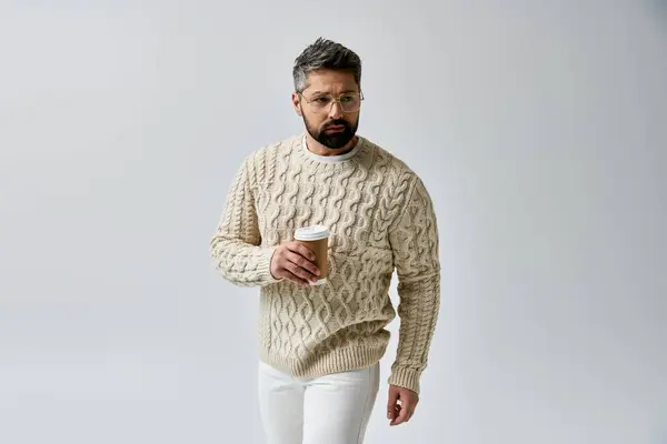 Un uomo accattivante con la barba che regge elegantemente una tazza di caffè, trasudando tranquillità e relax in un maglione bianco su uno sfondo grigio elegante. — Foto stock