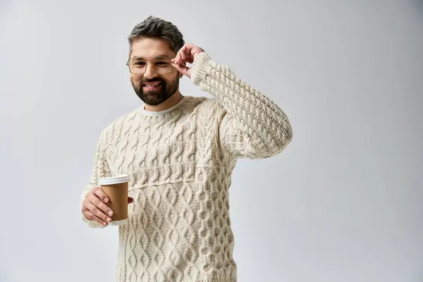 Un uomo barbuto con un maglione bianco tiene delicatamente una tazza di caffè fumante sullo sfondo di uno studio grigio. — Foto stock