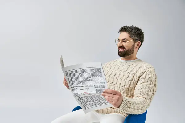 A stylish man with a beard sits on a chair, engrossed in reading a newspaper, against a grey background in a studio. — Stock Photo