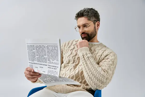 Un hombre barbudo en un suéter blanco se sienta en una silla, absorto en la lectura de un papel sobre un fondo gris estudio. - foto de stock