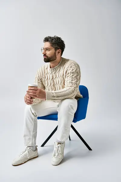 Um homem barbudo em uma camisola branca senta-se em uma cadeira azul, cruzando os braços em uma pose contemplativa contra um fundo cinza. — Fotografia de Stock