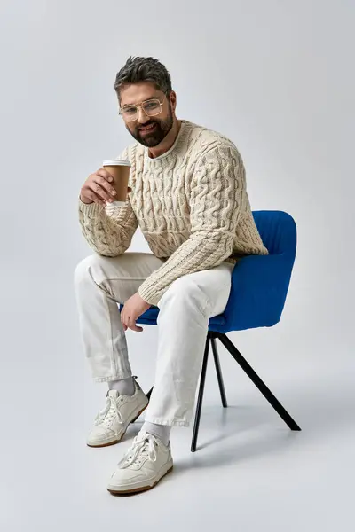A stylish man with a beard sits on a chair in a white sweater, savoring a cup of coffee on a grey studio backdrop. — Stock Photo