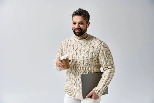 Ein bärtiger Mann im weißen Pullover hält zart eine Tasse Kaffee in der Hand und strahlt Wärme und Komfort in einer gemütlichen Studioatmosphäre aus.. — Stockfoto