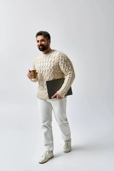 Бородатий чоловік у білому светрі тримає ноутбук і чашку кави, загубленої в думці. — стокове фото