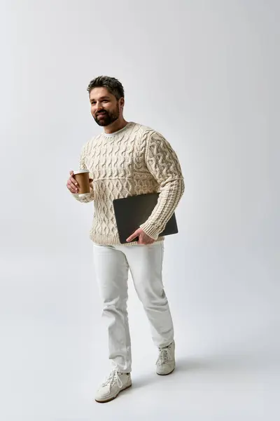 A bearded man in a white sweater holds a laptop and a cup of coffee, lost in thought. — Stock Photo