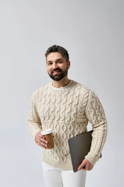 Ein bärtiger Mann im weißen Pullover hält eine Kaffeetasse und einen Laptop vor grauem Hintergrund. — Stockfoto