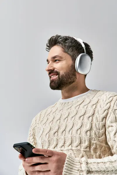 A bearded man in a white sweater wears headphones, engrossed in his cell phone screen against a grey backdrop. — Stock Photo
