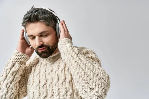 A stylish man with a beard wears a white sweater, immersed in music through headphones, against a grey studio backdrop. — Stock Photo