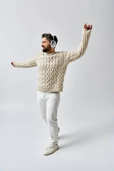 Ein stilvoller Mann mit Bart, Kopfhörer und weißem Pullover posiert vor grauem Studiohintergrund. — Stockfoto