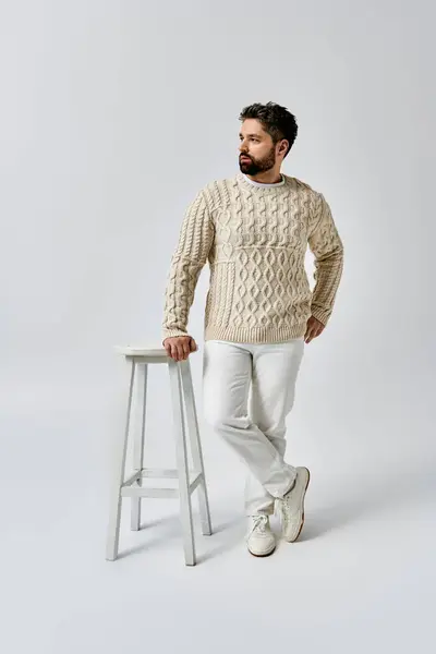 Ein stilvoller Mann mit Bart steht selbstbewusst neben einem Schemel und trägt einen modischen weißen Pullover in einem Studio-Ambiente. — Stockfoto