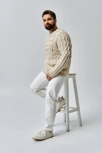 Ein bärtiger Mann versprüht Charme, als er auf einem Hocker sitzt, elegant gekleidet in einem weißen Pullover vor grauer Studiokulisse. — Stockfoto