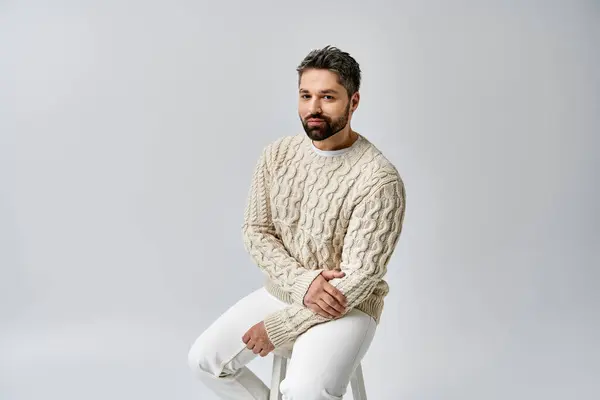 Un uomo carismatico con la barba si siede su uno sgabello, trasudando fascino in un accogliente maglione bianco sullo sfondo di uno studio grigio. — Foto stock