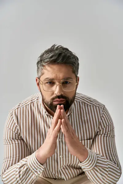 A stylish man with glasses and a beard sits gracefully, lost in thought on a grey background. — Stock Photo