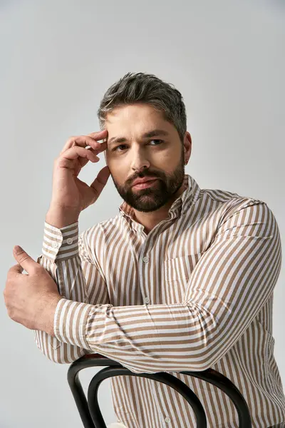 Um homem elegante com uma barba sentada em uma cadeira, exalando sofisticação e elegância, contra um fundo de estúdio cinza. — Stock Photo