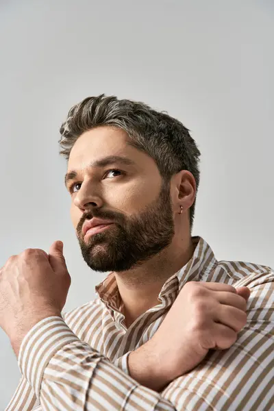 Um homem elegante com uma barba posa confiantemente em uma camisa listrada contra um fundo cinza. — Fotografia de Stock