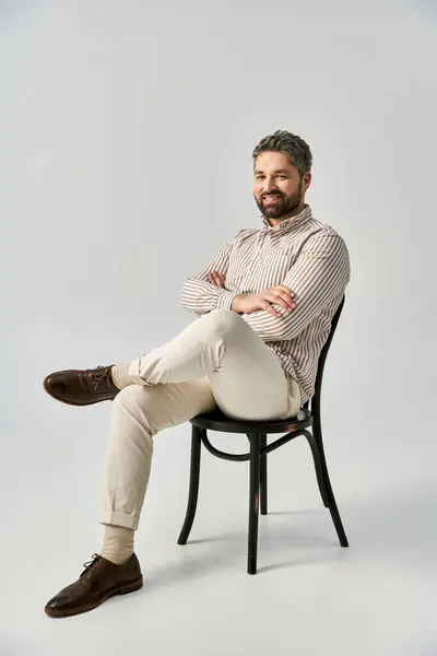 A bearded man in elegant attire sits on a chair with his legs crossed in a studio against a grey background. — Stock Photo