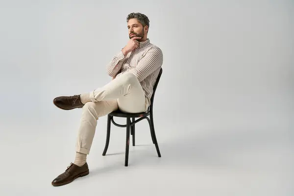 Стильний чоловік з бородою і елегантним вбранням сидить на стільці з упевненістю на сірому фоні студії. — стокове фото