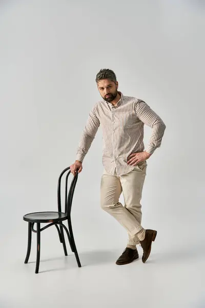 Un homme élégant avec une barbe se tient à côté d'une chaise, la main sur la hanche, respirant la confiance dans une tenue élégante sur un fond de studio gris. — Photo de stock
