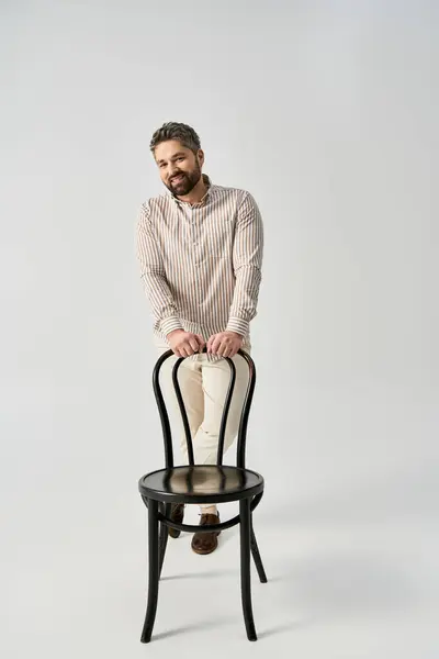A stylish man with a beard is sitting on top of a chair in an elegant pose against a grey studio background. — Stock Photo