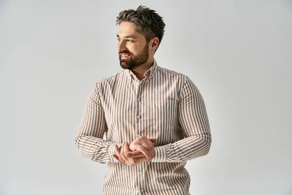 Un hombre barbudo está de pie con las manos dobladas, exudando elegancia y aplomo en atuendo de moda contra un fondo de estudio gris. - foto de stock