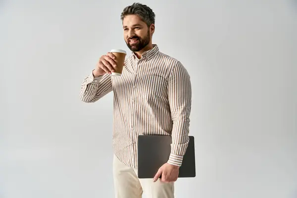 Ein stilvoller Mann in eleganter Kleidung mit Laptop und einer Tasse Kaffee vor grauem Hintergrund. — Stockfoto