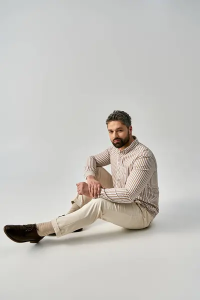 Un uomo barbuto in elegante abbigliamento seduto a terra con le gambe incrociate in una posa contemplativa su uno sfondo grigio studio. — Foto stock
