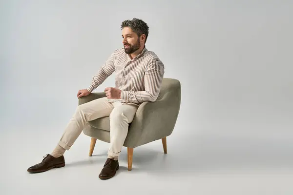 Homem barbudo em trajes elegantes senta-se com pernas cruzadas em pose elegante no fundo cinza no estúdio. — Fotografia de Stock