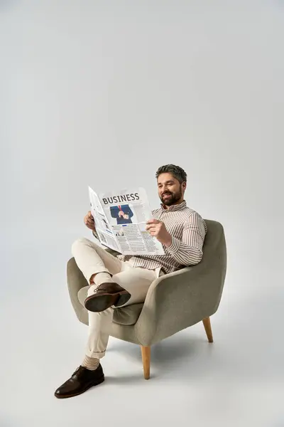 Un hombre elegante con barba se sienta en una silla, absorto en la lectura de un periódico. - foto de stock