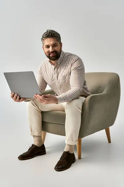 Стильний чоловік з бородою сидить на стільці, працюючи на своєму ноутбуці в модному і витонченому оточенні. — стокове фото