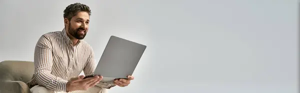 A bearded man in elegant attire sits on a chair, focused on his laptop screen. — Stock Photo