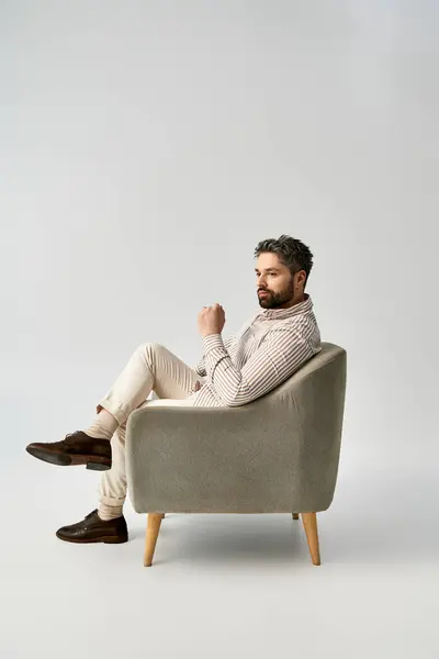 Un uomo barbuto in elegante abbigliamento si siede sulla poltrona su uno sfondo grigio studio. — Foto stock