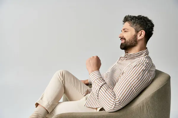 A stylish bearded man in elegant attire sits on armchair against a grey studio background. — Stock Photo