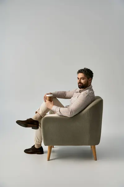 Un homme habillé avec une barbe se détend sur une chaise, savourant une tasse de café d'une manière élégante. — Photo de stock