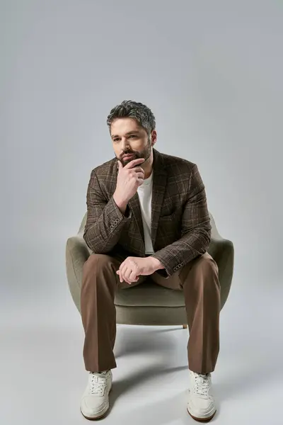 A bearded man in elegant attire sits with hand on chin, pondering deeply in a stylish pose against a grey studio background. — Stock Photo