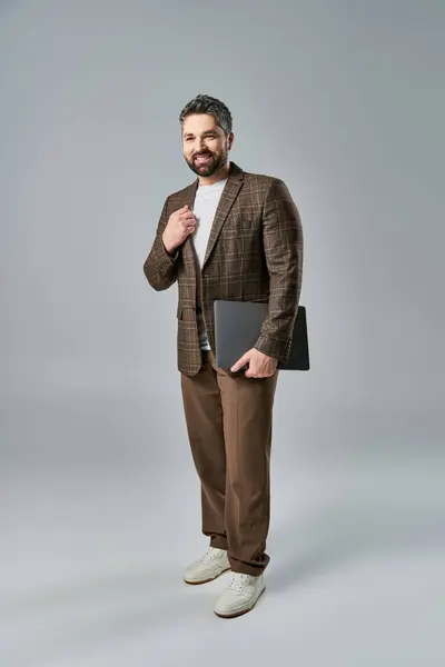 Бородатый мужчина в остром костюме, держащий ноутбук, излучающий профессионализм и изысканность на сером фоне студии. — стоковое фото