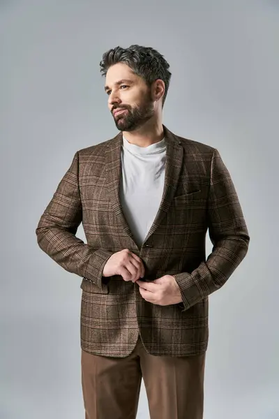 Un uomo carismatico con la barba che indossa una giacca marrone e pantaloni colpisce una posa in modo elegante su uno sfondo grigio studio. — Stock Photo
