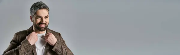 A bearded man strikes an alluring pose in an elegant brown jacket and white t-shirt on a grey studio background. — Stock Photo