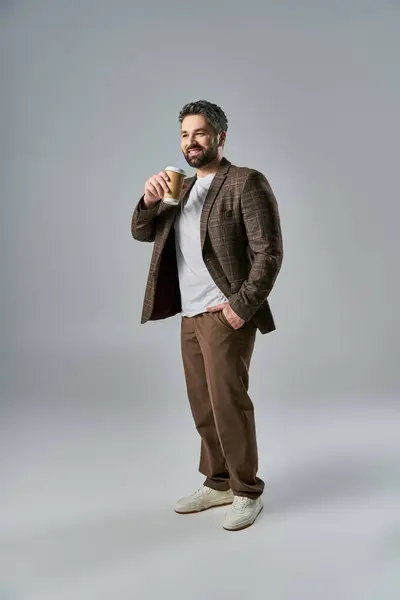 Selbstbewusst steht ein stilvoller Mann mit Bart vor grauem Hintergrund und hält eine dampfende Tasse Kaffee in der Hand.. — Stockfoto