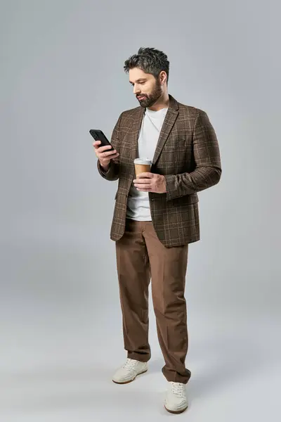 Стильный мужчина в элегантной одежде, держащий чашку кофе и проверяющий телефон, затерявшись в мыслях. — стоковое фото