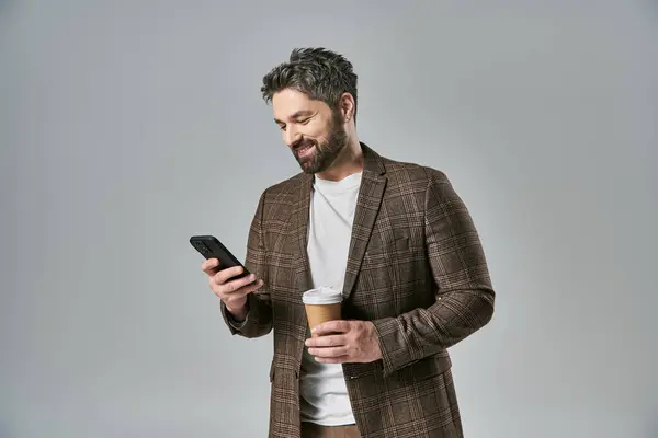 Ein kultivierter Mann mit Bart in eleganter Kleidung hält eine Tasse Kaffee in der Hand, während er auf sein Handy blickt. — Stockfoto