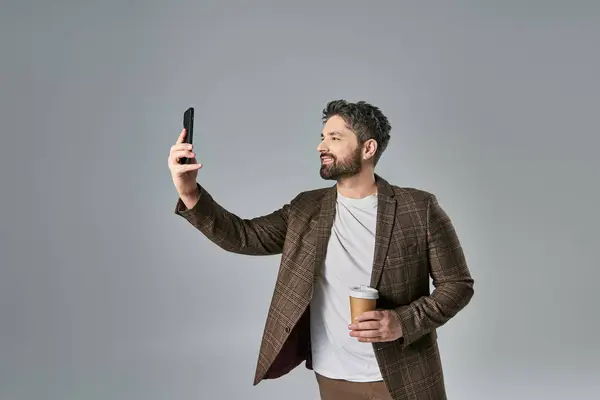 Un homme élégant avec une barbe vêtue d'une tenue élégante prenant un autoportrait avec son téléphone portable sur un fond gris. — Photo de stock